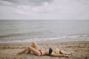 Chica en forma con sombrero acostada en la playa. Mujer joven de moda que cubre con sombrero de paja, relajándose en la playa de arena cerca del mar. Vacaciones de verano y viajes. Mindfulness y despreocupación