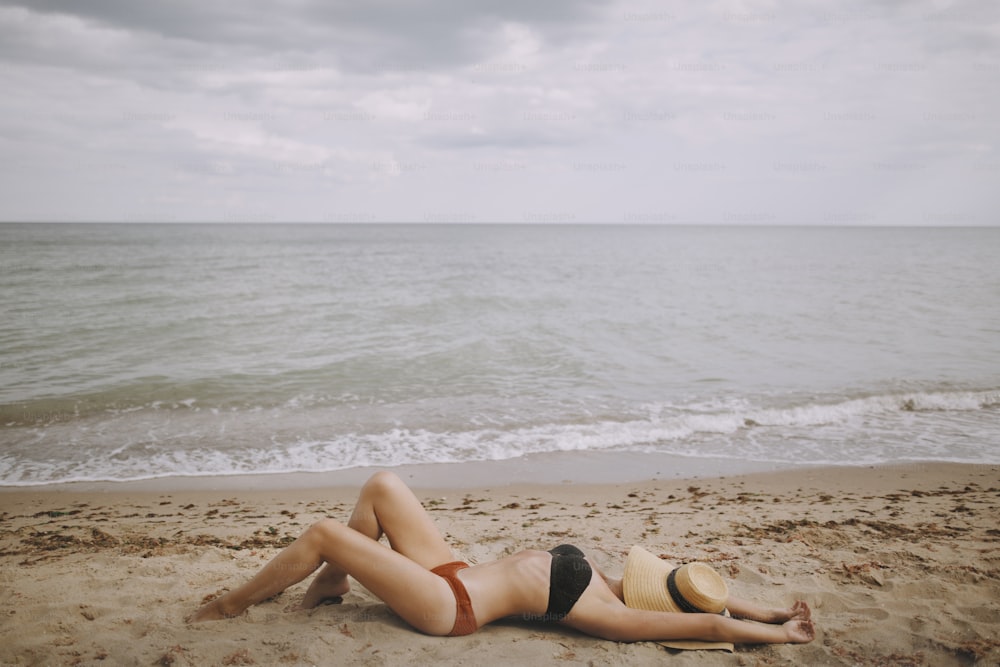 Fittes Mädchen mit Hut am Strand liegend. Modische junge Frau mit Strohhut, entspannt am Sandstrand in der Nähe des Meeres. Sommerurlaub und Reisen. Achtsamkeit und Sorglosigkeit