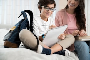 Smiling focused schoolboy in eyeglasses doing his homework in the presence of his female parent