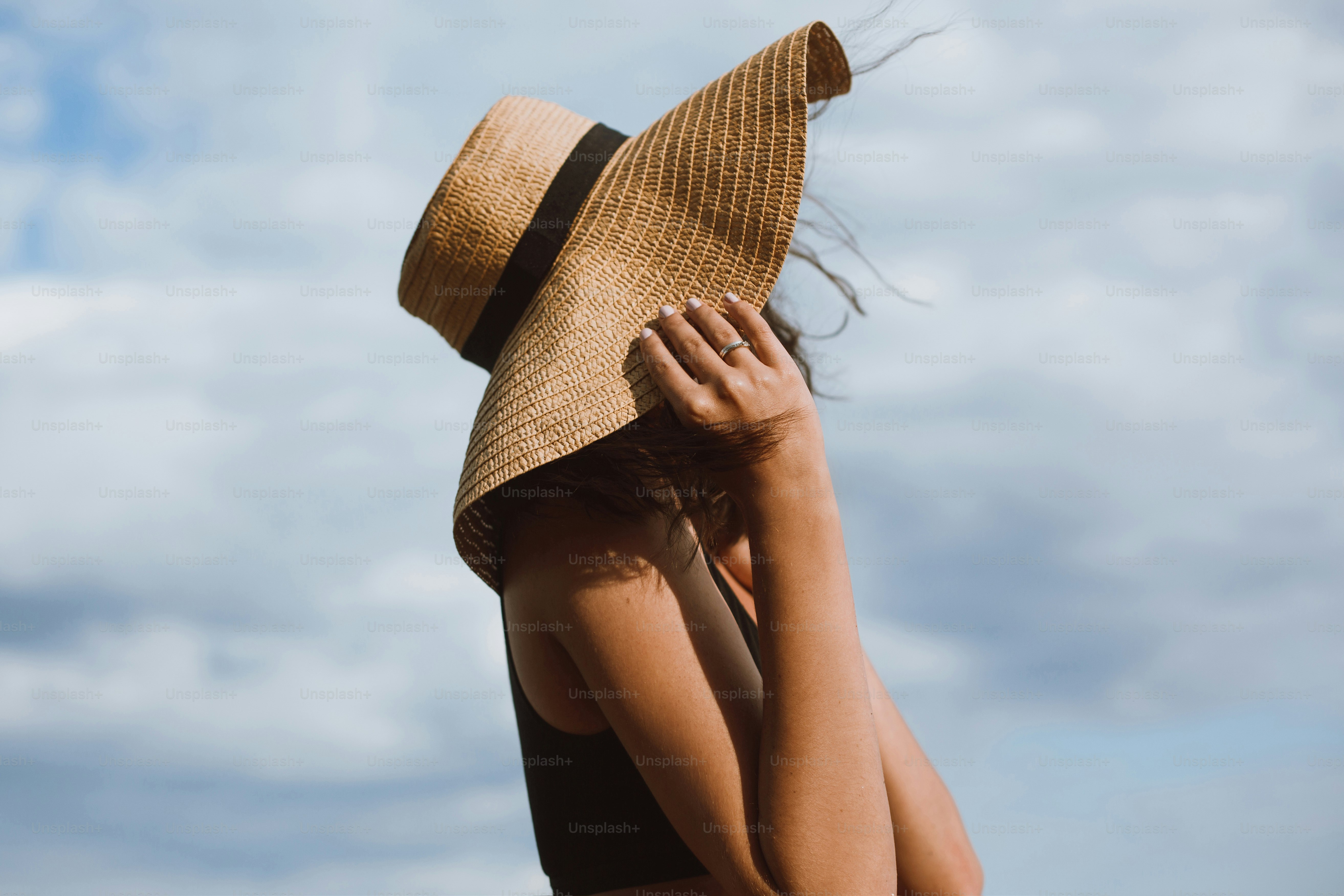 Chica bronceada sosteniendo sombrero y posando sobre fondo de cielo azul en un caluroso día de verano. Mujer joven de moda con sombrero de paja relajándose en la playa. Despreocupado. Veraneo
