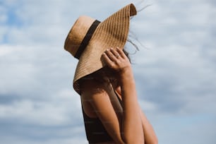 Tanned girl holding hat and posing on background of blue sky in hot summer day. Fashionable young woman in straw hat relaxing on beach. Carefree. Summer vacation