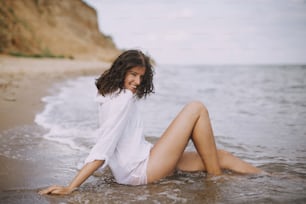 Mulher jovem feliz na camisa branca sentada na praia em ondas espirrando. Elegante menina bronzeada relaxando à beira-mar e desfrutando de ondas. Férias de verão. Mindfulness e momento despreocupado