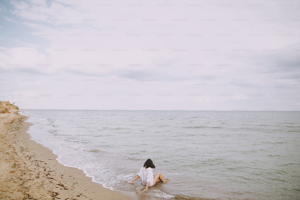 波しぶきをあげるビーチに座っている白いシャツを着た幸せな若い女性。海岸でリラックスし、波を楽しむスタイリッシュな日焼けした女の子。夏休み。マインドフルネスと気楽なひととき