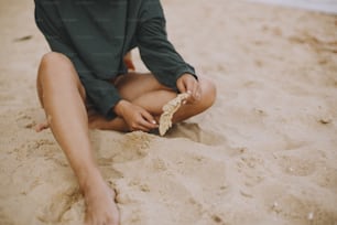 Chica hipster despreocupada sosteniendo hierba y sentada en la playa, vista recortada en piernas bronceadas. Elegante mujer boho en traje de baño moderno y suéter relajándose en la orilla del mar. Veraneo. Atención plena