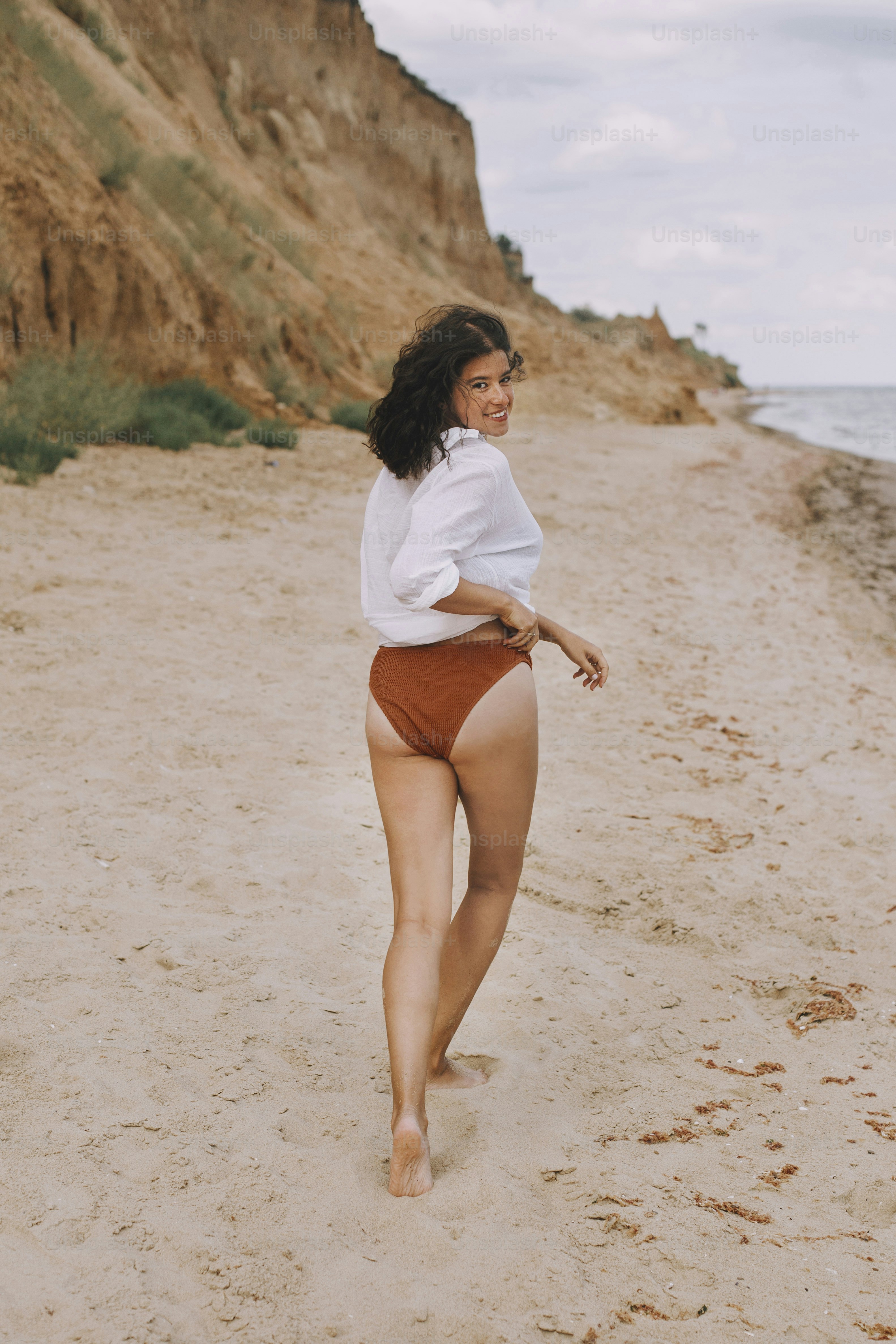 Happy boho girl in white shirt running on sunny beach. Carefree stylish woman in swimsuit and shirt relaxing on seashore. Summer vacation. Lifestyle authentic image