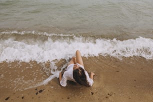 Young woman in wet white shirt lying on beach in splashing waves. Top view. Stylish tanned girl relaxing on seashore and enjoying waves. Summer vacation. Mindfulness and carefree