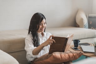 Mulher asiática jovem feliz lendo livro sentado no sofá em casa.