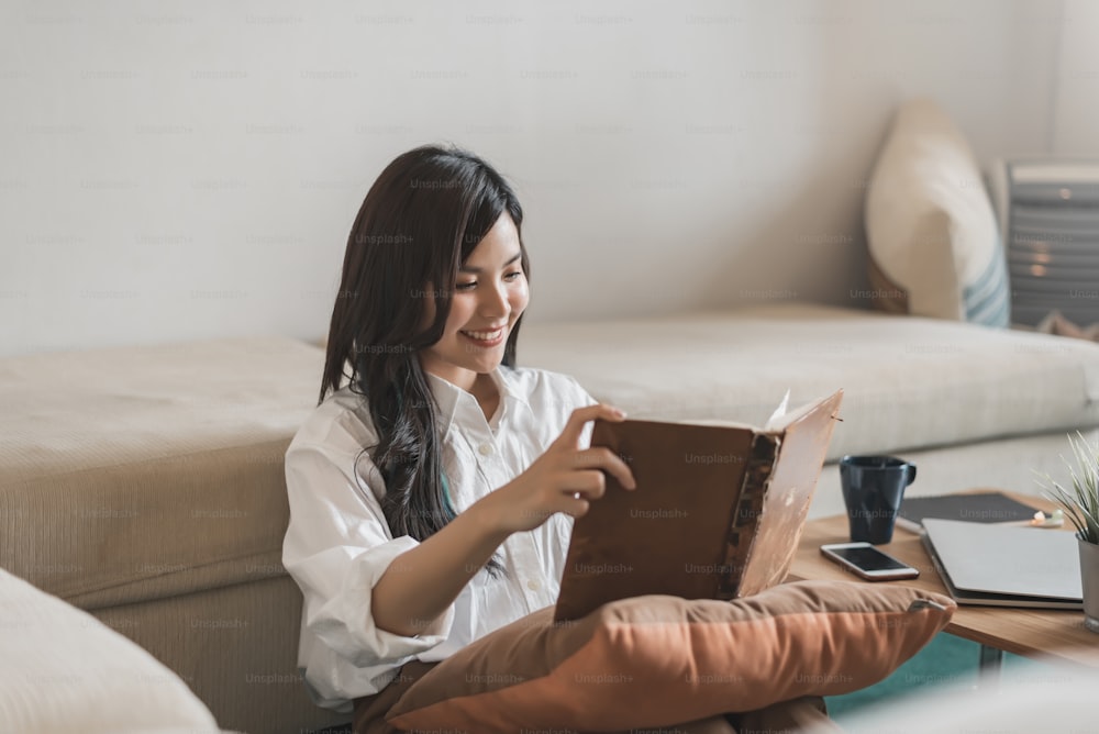 Happy young asian woman reading book sitting on the sofa at home.
