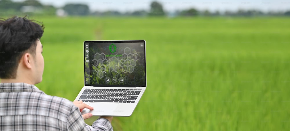 Cropped image of young smart farmer holding a computer laptop with visual icon on screen over rice field as background. Agriculture technology concept.