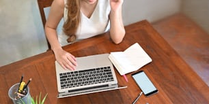 Top view image of stylish woman in sexy dress typing on computer laptop that putting on wooden working desk that surrounded by notebook, white screen mobile, pencil holder and potted plant.