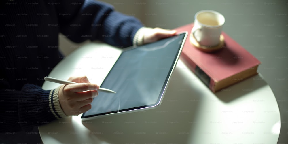 Cropped shot of a woman writing on mock-up tablet while sitting at white coffee table with book and coffee cup