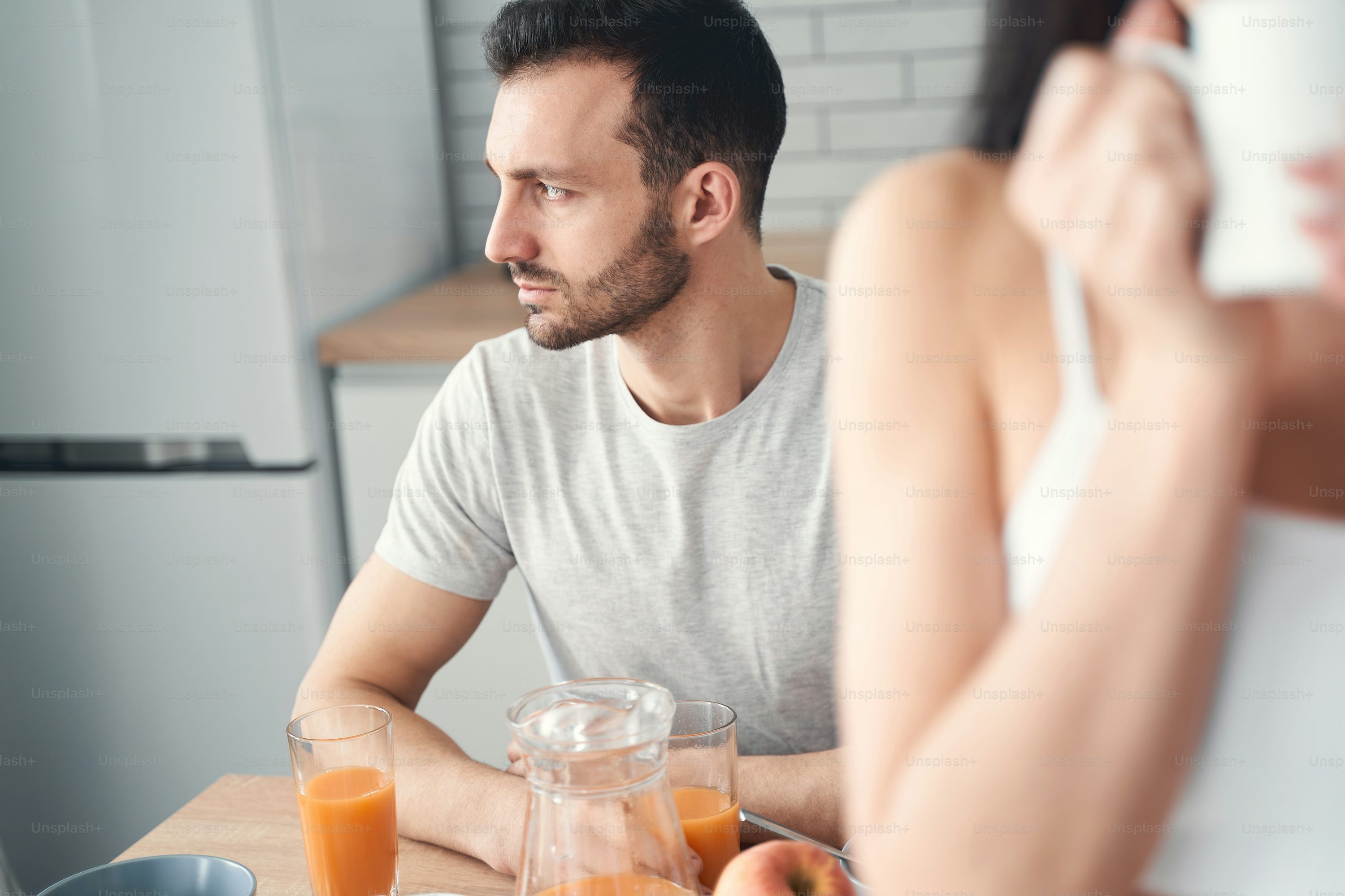 Handsome bearded male sitting at kitchen table with glass of fresh juice in hands while looking aside thoughtfully