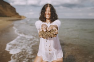 Happy young woman in white shirt holding wet sand in hands on beach. Funny tanned girl relaxing on seashore. Summer vacation. Carefree funny moment
