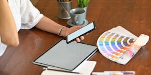 Closeup graphic designer holding/using a white blank screen smartphone while sitting at wooden working desk that surrounded by empty screen computer tablet, color guide and office equipment.
