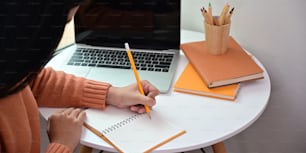Abgeschnittenes Bild einer attraktiven Frau, die Notizen macht, während sie am weißen Schreibtisch über einem gemütlichen Wohnzimmer als Hintergrund sitzt.