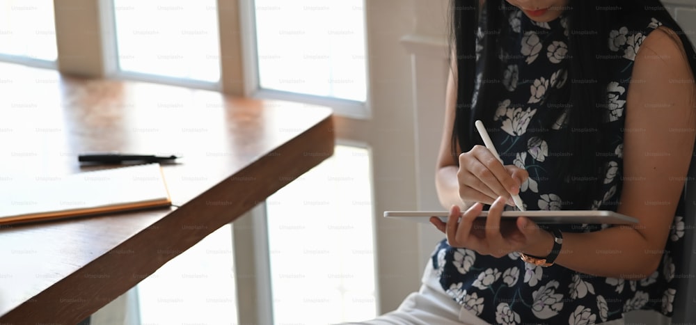 Photo of beautiful woman drawing/sketching on computer tablet by using a stylus pen while sitting at the working desk over comfortable sitting room as background.