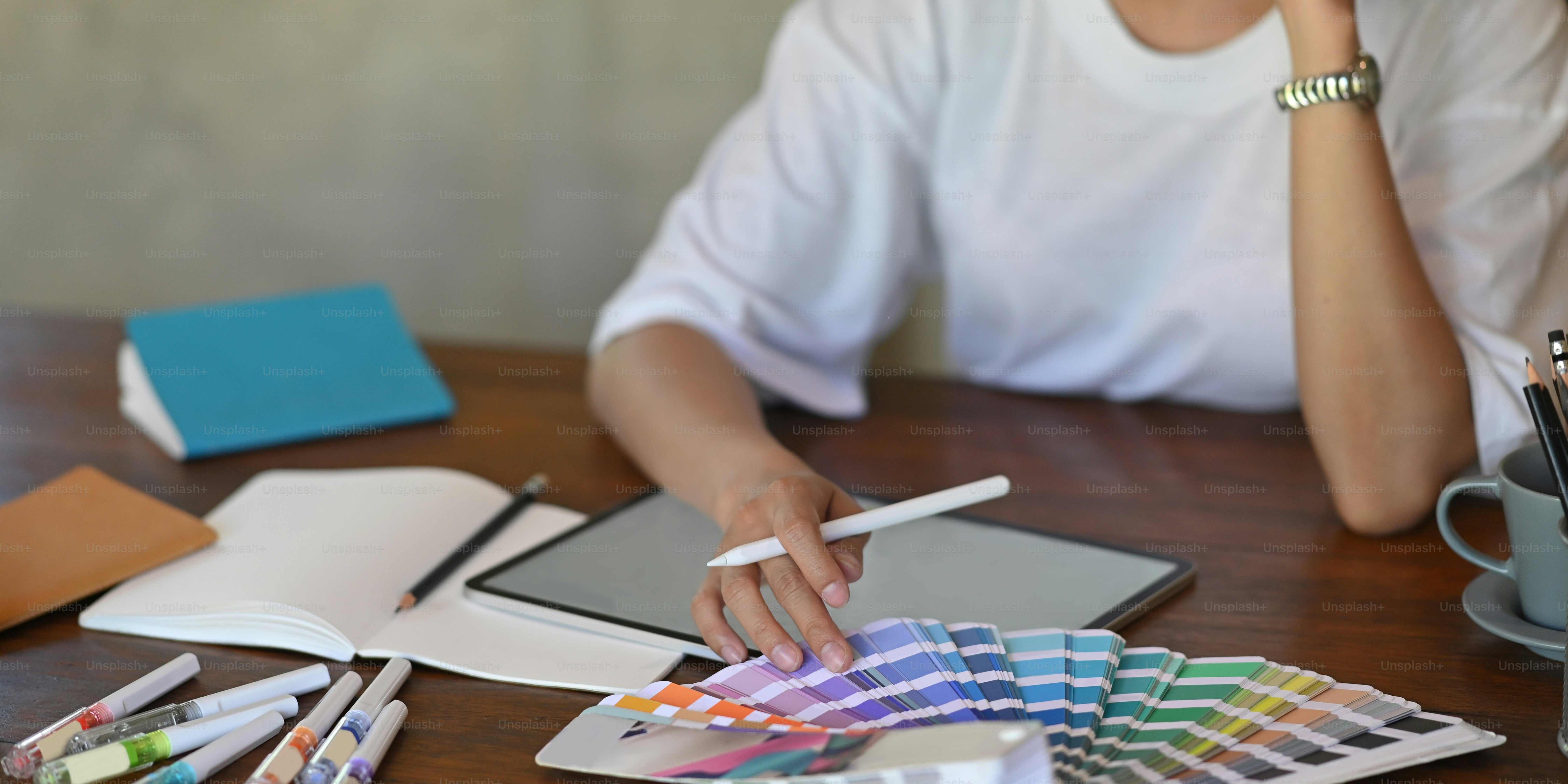 Photo of young graphic designer holding a stylus pen while selecting color from color guide and sitting in front of computer tablet with empty screen that putting on wooden working desk.