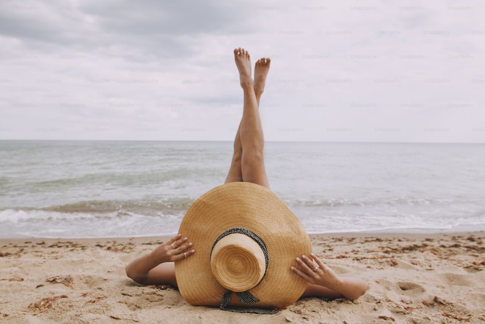 Chica con sombrero acostada en la playa con las piernas levantadas. Mujer joven de moda que cubre con sombrero de paja, relajándose en la playa de arena cerca del mar. Vacaciones de verano y viajes. Mindfulness y despreocupación