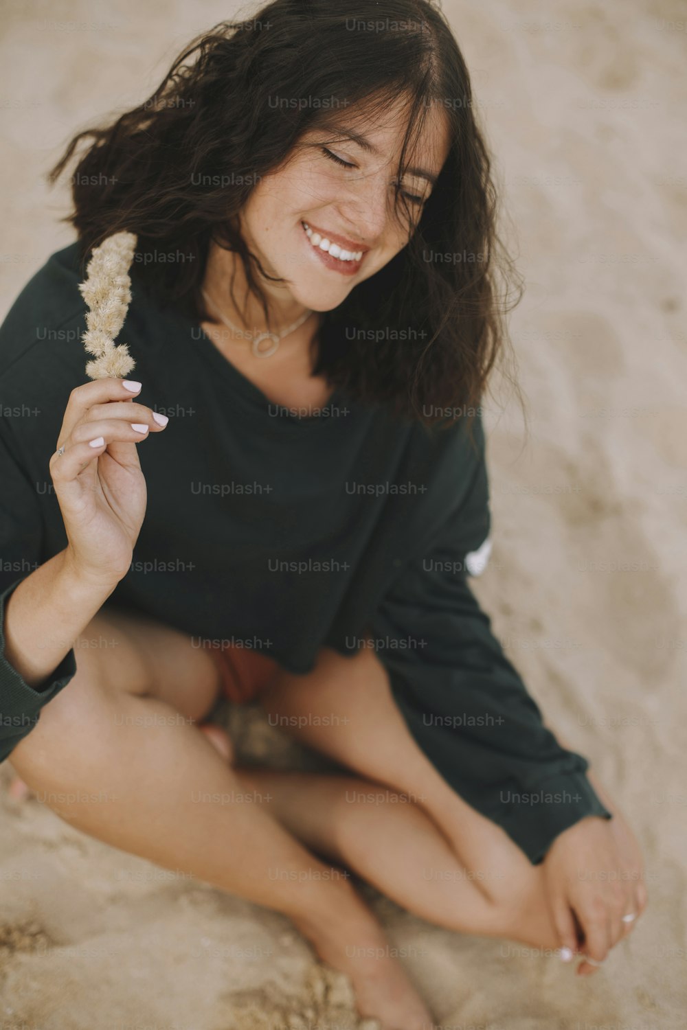 Menina hipster despreocupada com cabelo ventoso sentado e sorrindo na praia de areia, segurando erva. Elegante mulher jovem bronzeada em maiô moderno e suéter relaxando à beira-mar. Férias de verão