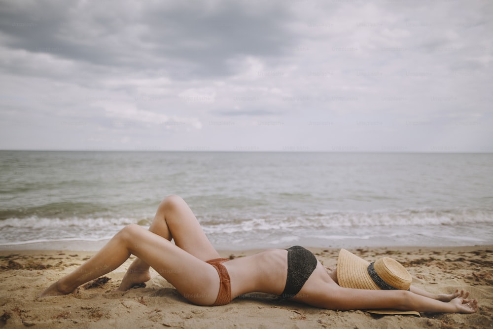 Tanned girl in hat lying on beach. Fashionable young woman covering with straw hat, relaxing on sandy beach near sea. Summer vacation and travel. Mindfulness and carefree