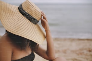 Girl sitting on beach, hand holding hat close up. Fashionable young woman in straw hat relaxing on sandy beach near sea. Carefree. Space for text. Summer vacation and travel.
