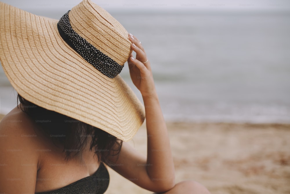 Girl sitting on beach, hand holding hat close up. Fashionable young woman in straw hat relaxing on sandy beach near sea. Carefree. Space for text. Summer vacation and travel.