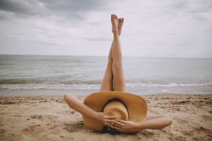 Girl in hat lying on beach with legs up. Fashionable young woman covering with straw hat, relaxing on sandy beach near sea. Summer vacation and travel. Mindfulness and carefree