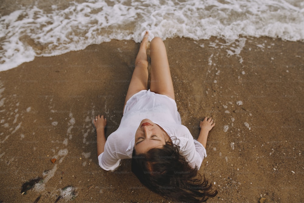濡れた白いシャツを着た若い女性が波しぶきを上げてビーチに横たわっています。上面図。海岸でリラックスし、波を楽しむスタイリッシュな日焼けした女の子。夏休み。マインドフルネスと気楽