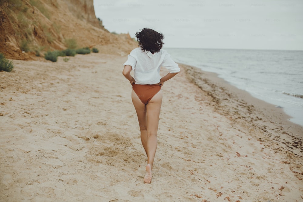 햇볕이 잘 드는 해변에서 달리는 흰 셔츠를 입은 보헤미안 소녀, 뒷모습. 수영복과 셔츠를 입고 해변에서 휴식을 취하는 평온한 세련된 여성. 여름 방학. 라이프 스타일 정통 이미지