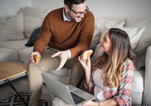 J’ai trouvé quelque chose qui va vous intéresser. Couple heureux à la maison utilisant un ordinateur portable parlant et mangeant un sandwich.