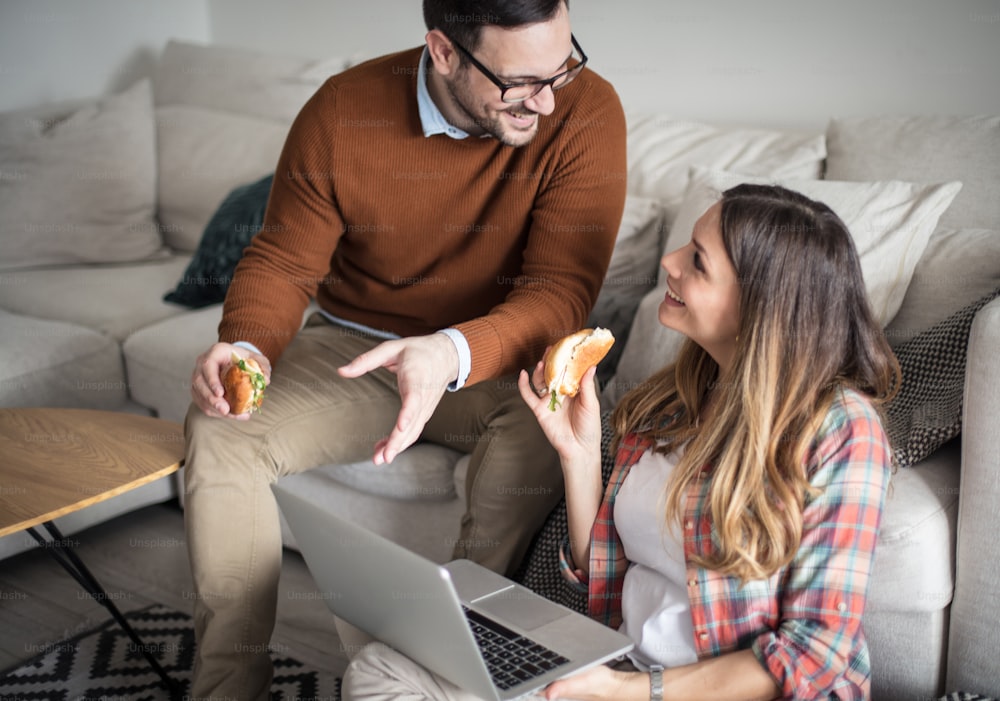 I found something that will interest you. Happy couple at home using laptop talking and eating sandwich.