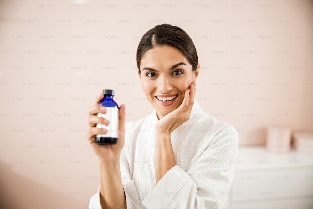 Portrait of cheerful beautiful lady in bathrobe holding bottle of remedy for health and care isolated on pink background