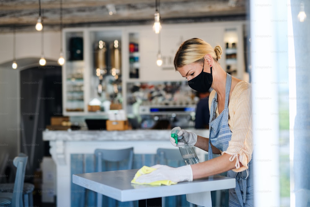 Mujer propietaria de una cafetería trabajando con mascarilla y guantes, limpiando y desinfectando mesas.