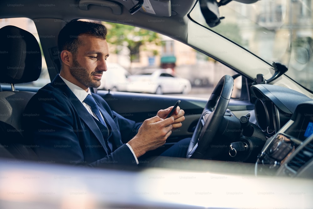 Handsome Caucasian businessman in formal clothes spending time in auto while looking on smartphone