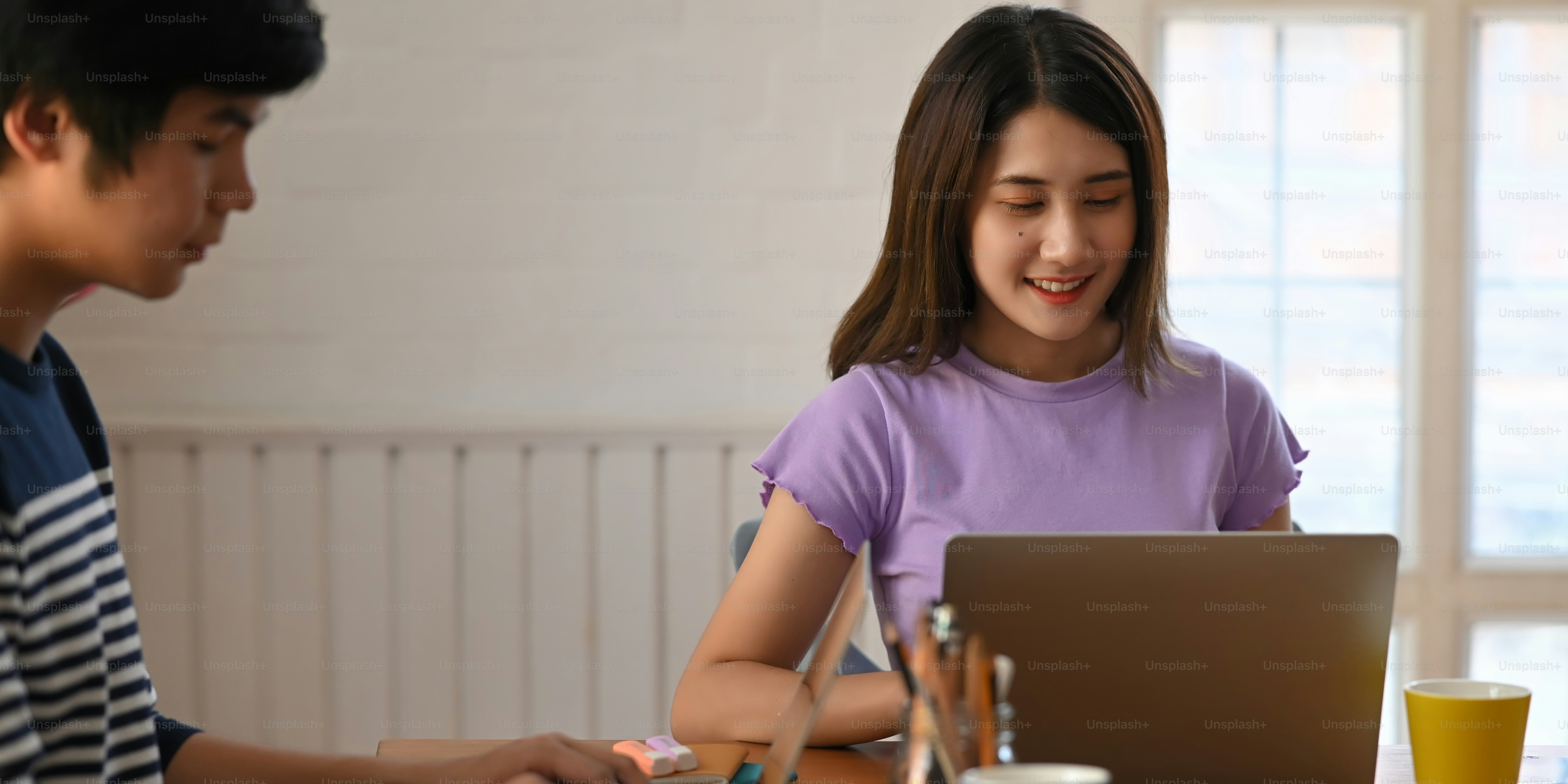 Pasangan muda yang bekerja dengan komputer, laptop, dan tablet sambil duduk bersama di meja kerja kayu di atas ruang duduk yang nyaman sebagai latar belakang.