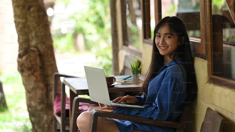 Eine schöne Frau benutzt einen Computer-Laptop, während sie auf dem Holzstuhl sitzt.