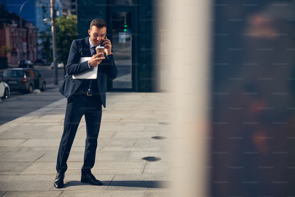 Handsome brunette businessman holding device and coffee in hands while speaking on phone and smiling
