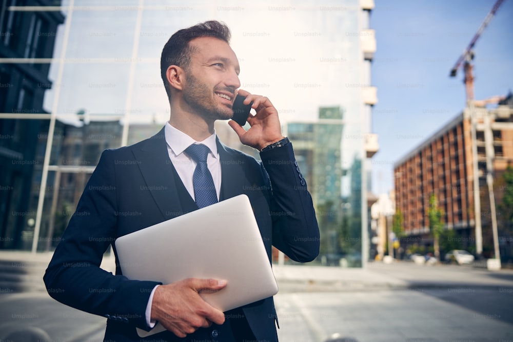 Successful Caucasian man in beautiful suit looking happily while speaking on smartphone outdoors and smiling