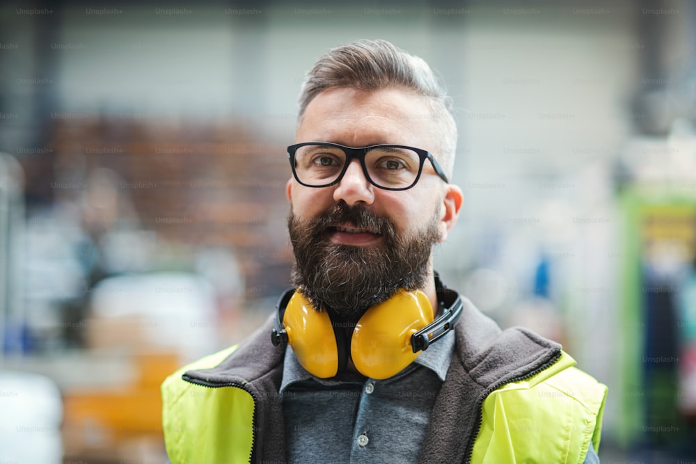Technician or engineer with protective headphones standing in industrial factory, looking at camera.