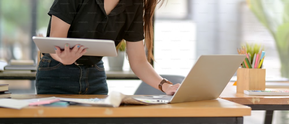 Colpo ritagliato di designer donna che lavora con tablet, laptop e forniture di design mentre si trova al tavolo da lavoro