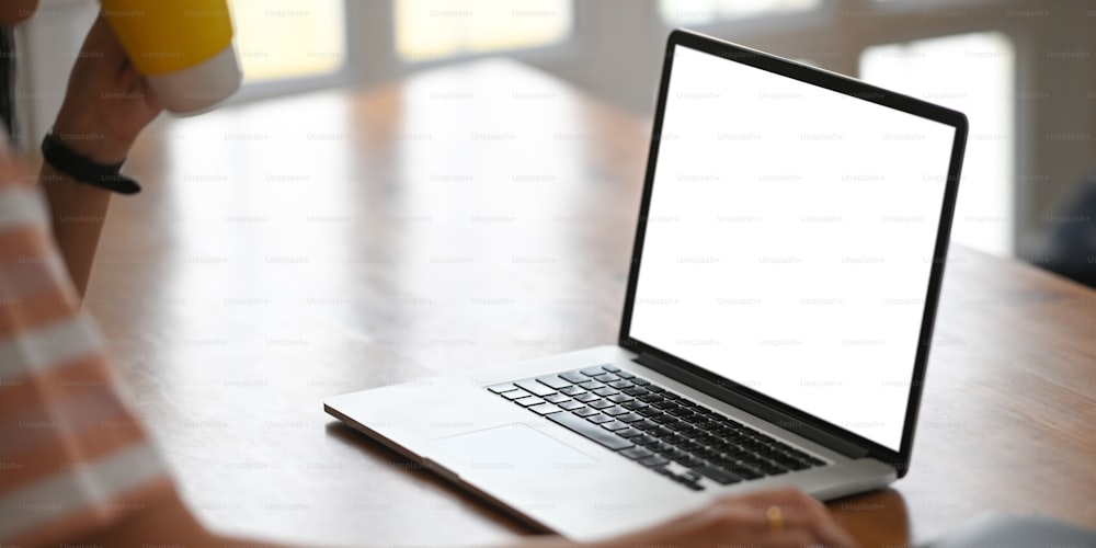 A beautiful woman is drinking coffee while working on a white blank screen computer laptop that putting on a wooden working desk.