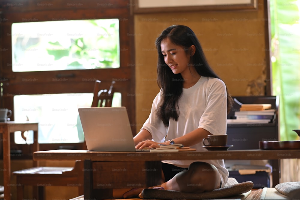 아름다운 여자가 나무 짧은 다리 테이블에서 컴퓨터 노트북에 타이핑하고 있습니다.