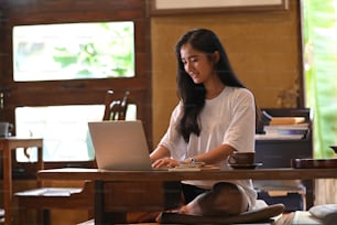 A beautiful woman is typing on a computer laptop at the wooden short legs table.