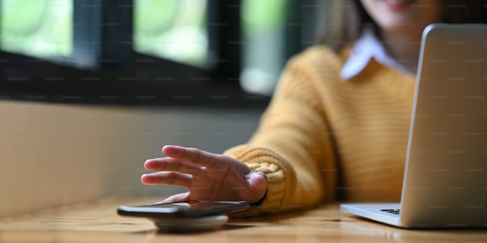 Image recadrée d’une femme de bureau chargeant un smartphone avec un chargeur sans fil tout en tapant sur un ordinateur portable.