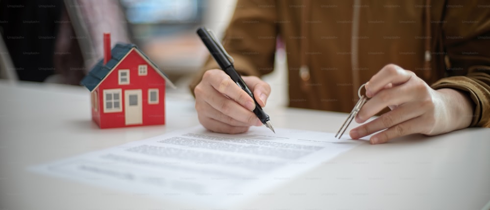 Abgeschnittene Aufnahme einer Frau, die einen Wohnungsbaudarlehensvertrag unterzeichnet, während sie den Hausschlüssel auf einem weißen Tisch mit Hausmodell hält