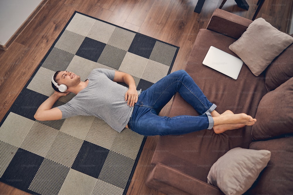 Hanfsome young male staying alone on floor with legs on couch while enjoying time in headphones