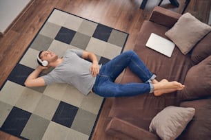 Hanfsome joven masculino que se queda solo en el suelo con las piernas en el sofá mientras disfruta del tiempo con auriculares