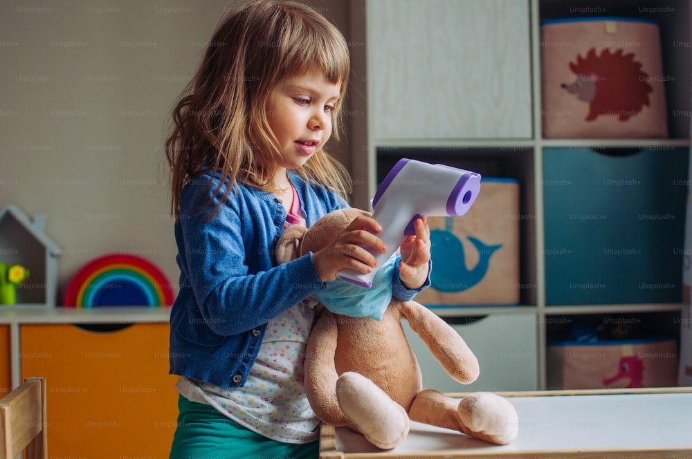 Toddler girl using electronic non-contact thermometer for bunny soft toy in the kids room. Pandemic COVID-19 concept.