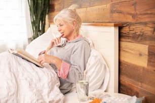Saisonale Grippe. Kranke ältere Frau liest Buch im Bett, zu Hause.