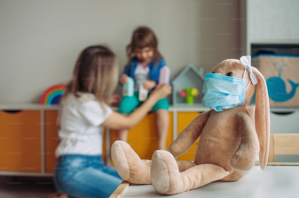 Bunny soft toy in the medicine mask in the kids room. Defocused happy family mother and daughter playing together as a background. Pandemic COVID-19 concept.